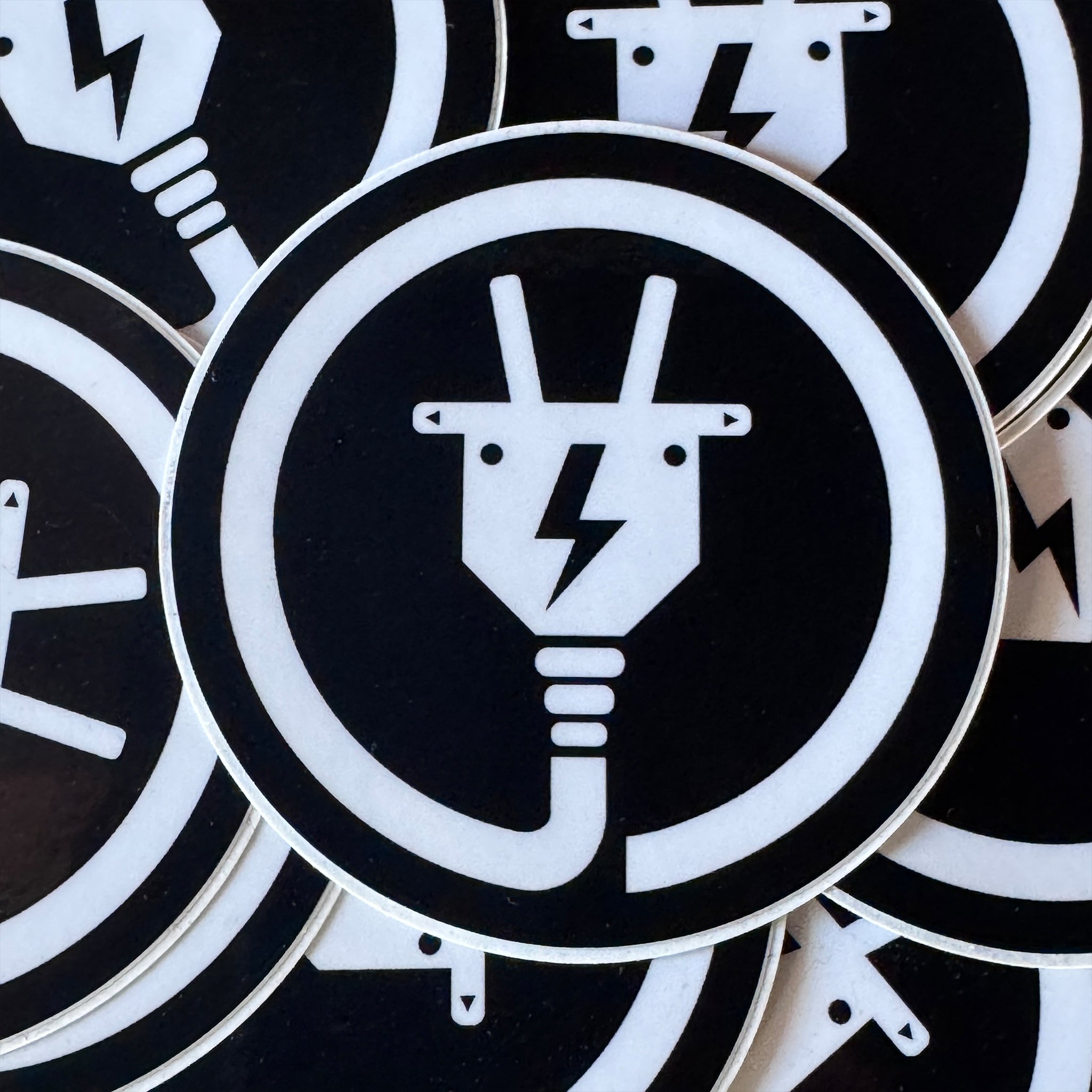 Circular black and white symbol from The Baby Goats with Electricity plug design on a black background.